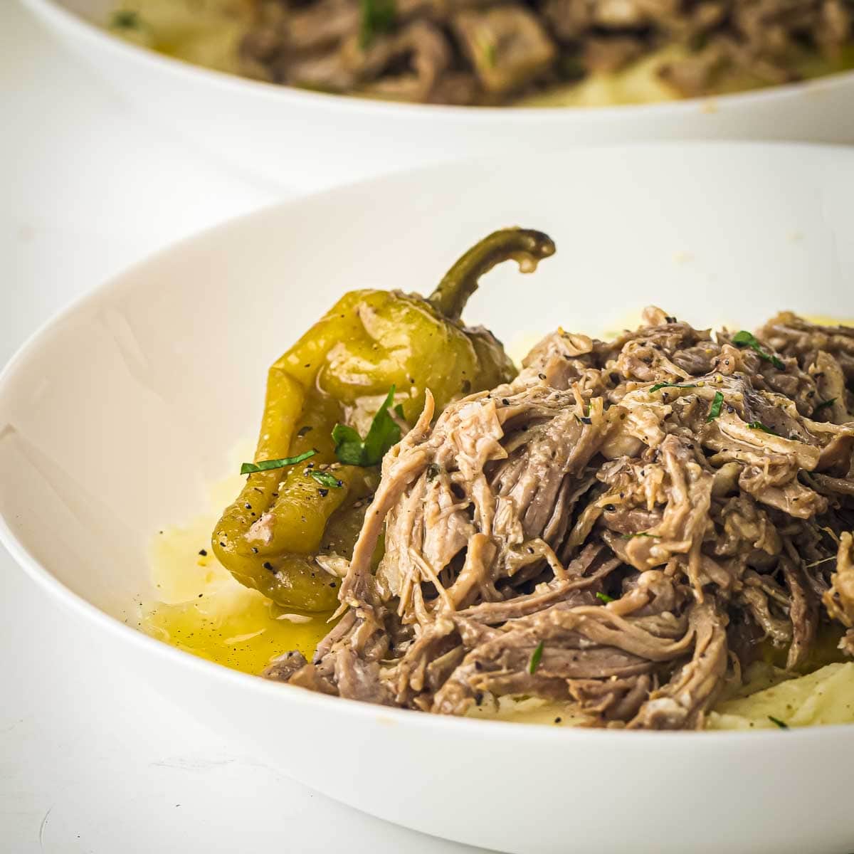 Side shot of pork roast on top of mashed potatoes in a white bowl with a pepperoncini on the side and garnished with fresh herbs.