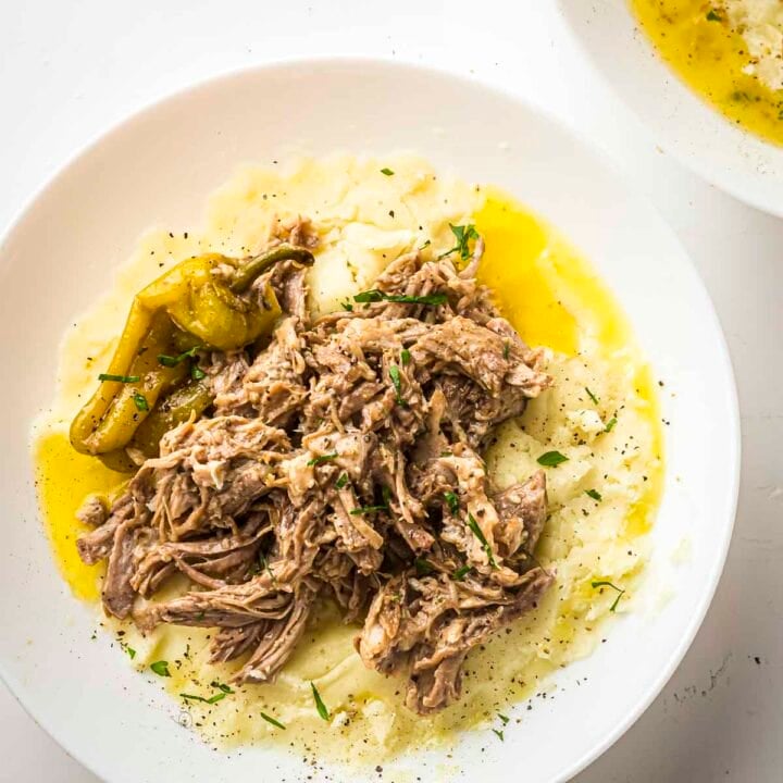 Overhead view of pork roast on top of mashed potatoes in a white bowl with a pepperoncini on the side and garnished with fresh herbs.