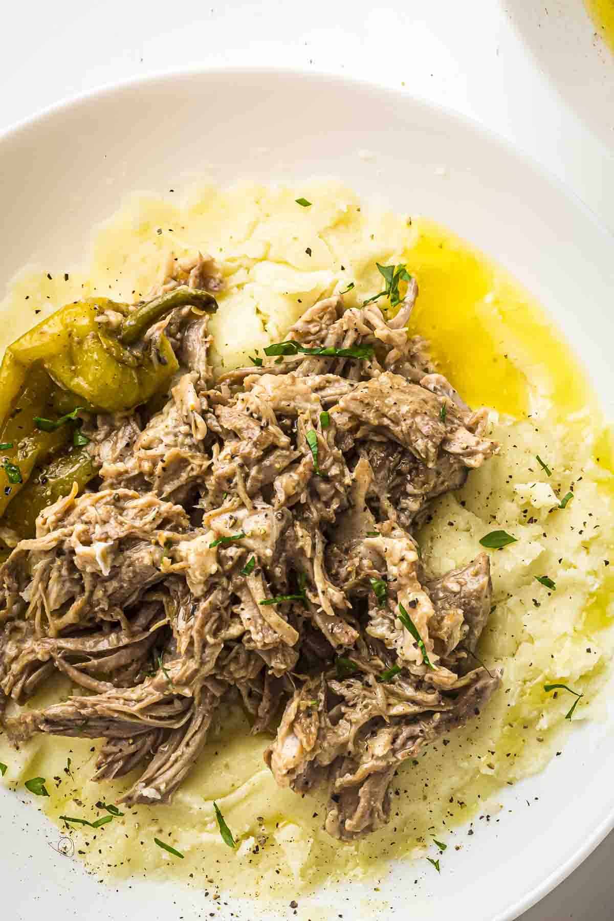 Overhead view of pork roast on top of mashed potatoes in a white bowl with a pepperoncini on the side and garnished with fresh herbs.