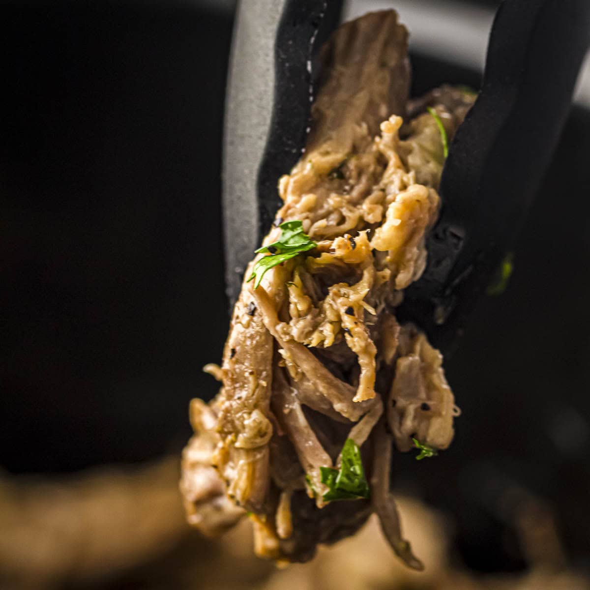 Close up shot of tongs holding up a piece of slow cooker pork roast.