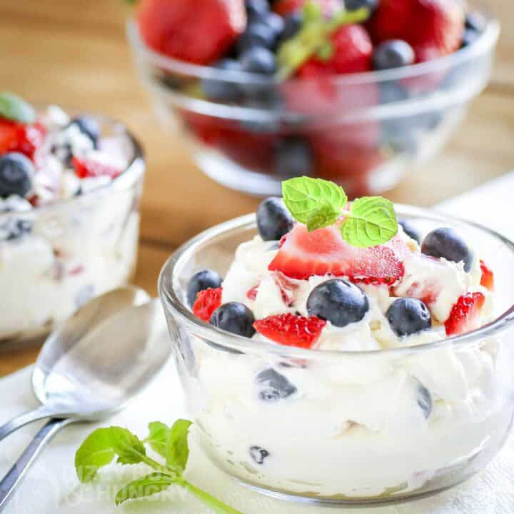 Side shot of a small glass bowl filled with cheesecake fluff topped with fresh blueberries and strawberries and fresh basil with more fresh fruits in the background.