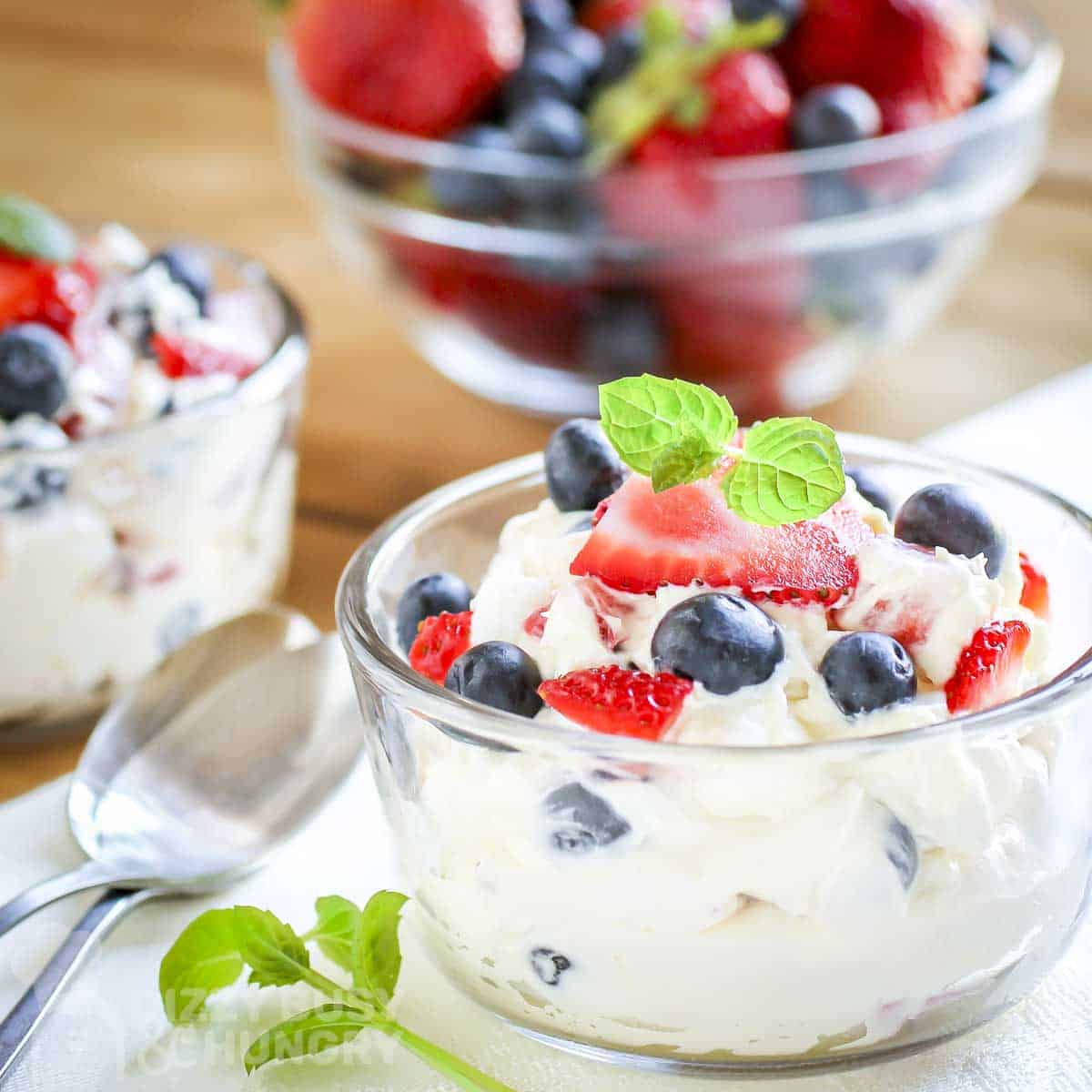 Side shot of a small glass bowl filled with cheesecake fluff topped with fresh blueberries and strawberries and fresh basil with more fresh fruits in the background.