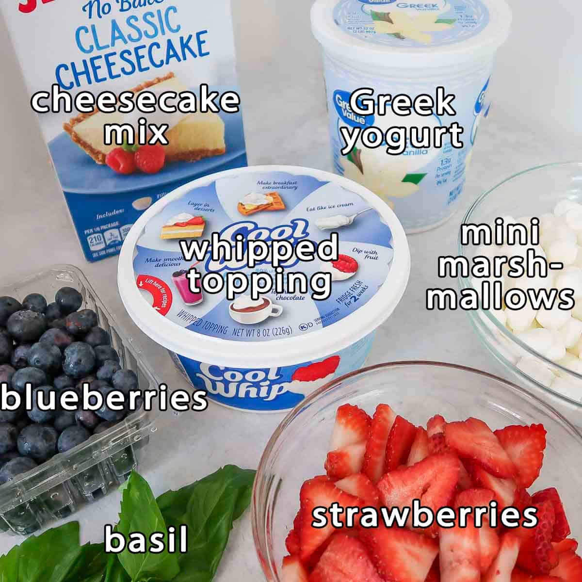 Side shot of ingredients - cheesecake mix, greek yogurt, whipped topping, mini marshmallows, blueberries, basil, and strawberries.