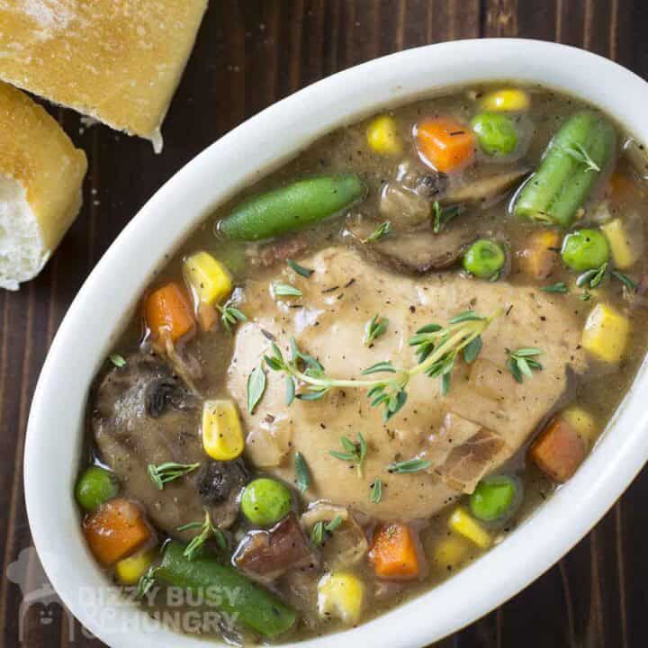 Overhead shot of chicken stew garnished with fresh thyme in a white oval bowl on a wooden surface with sliced bread on the side.