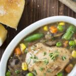 Overhead shot of chicken stew garnished with fresh thyme in a white oval bowl on a wooden surface with sliced bread on the side.