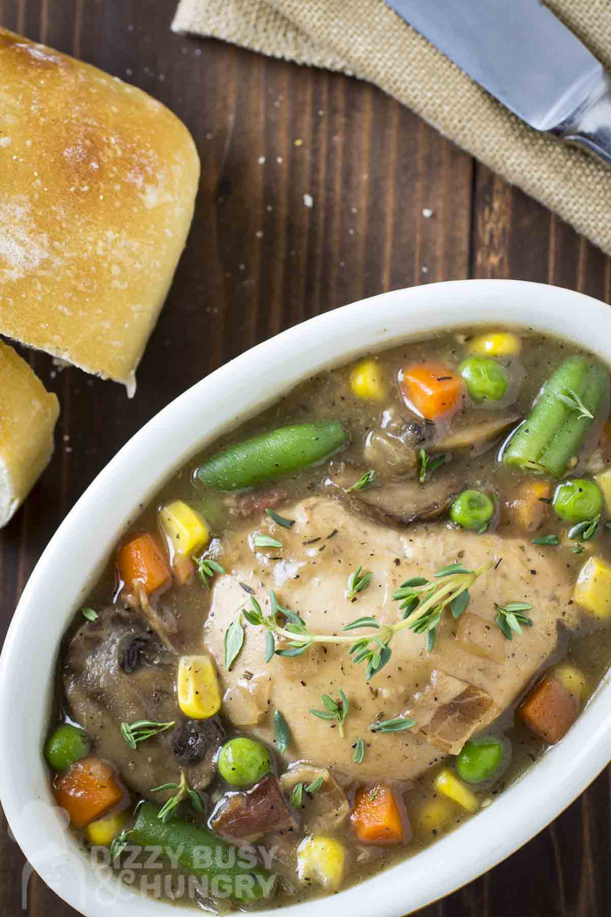 Overhead shot of chicken stew garnished with fresh thyme in a white oval bowl on a wooden surface with sliced bread on the side.