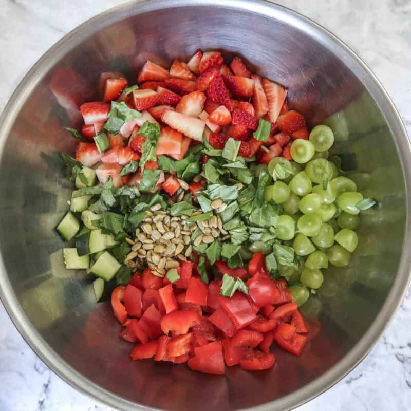 Overhead view of process shot 1 - chop the cucumber, strawberries, grapes, basil leaves, and red bell pepper into bite sized chunks. Place everything in a large bowl and add in the sunflower seeds.