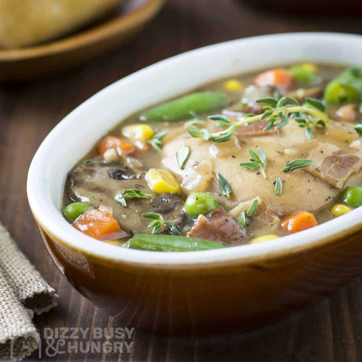 Side shot of chicken stew garnished with fresh thyme in a white oval bowl on a wooden surface with sliced bread on the side.