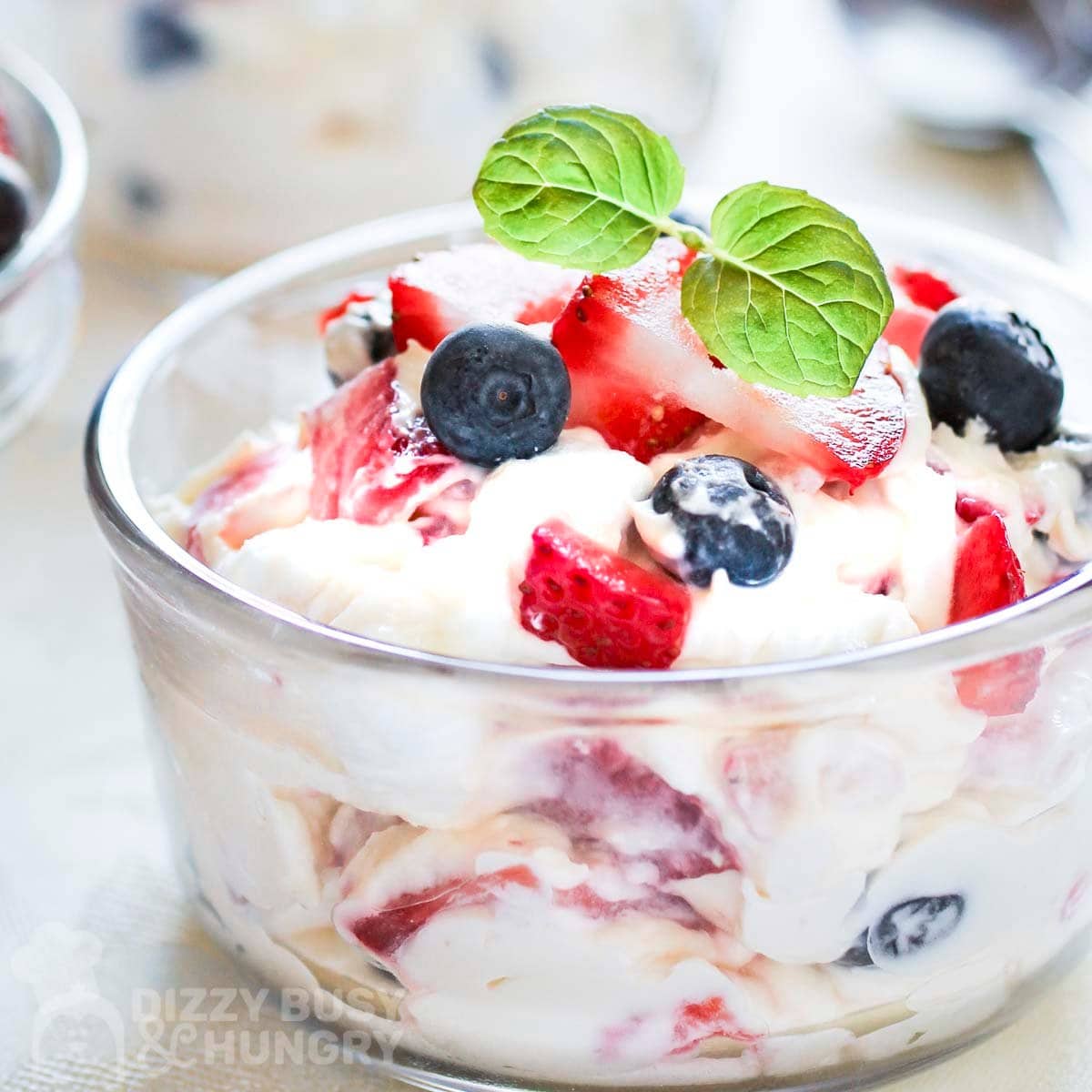 Side shot of a small glass bowl filled with cheesecake fluff topped with fresh blueberries and strawberries and fresh basil.