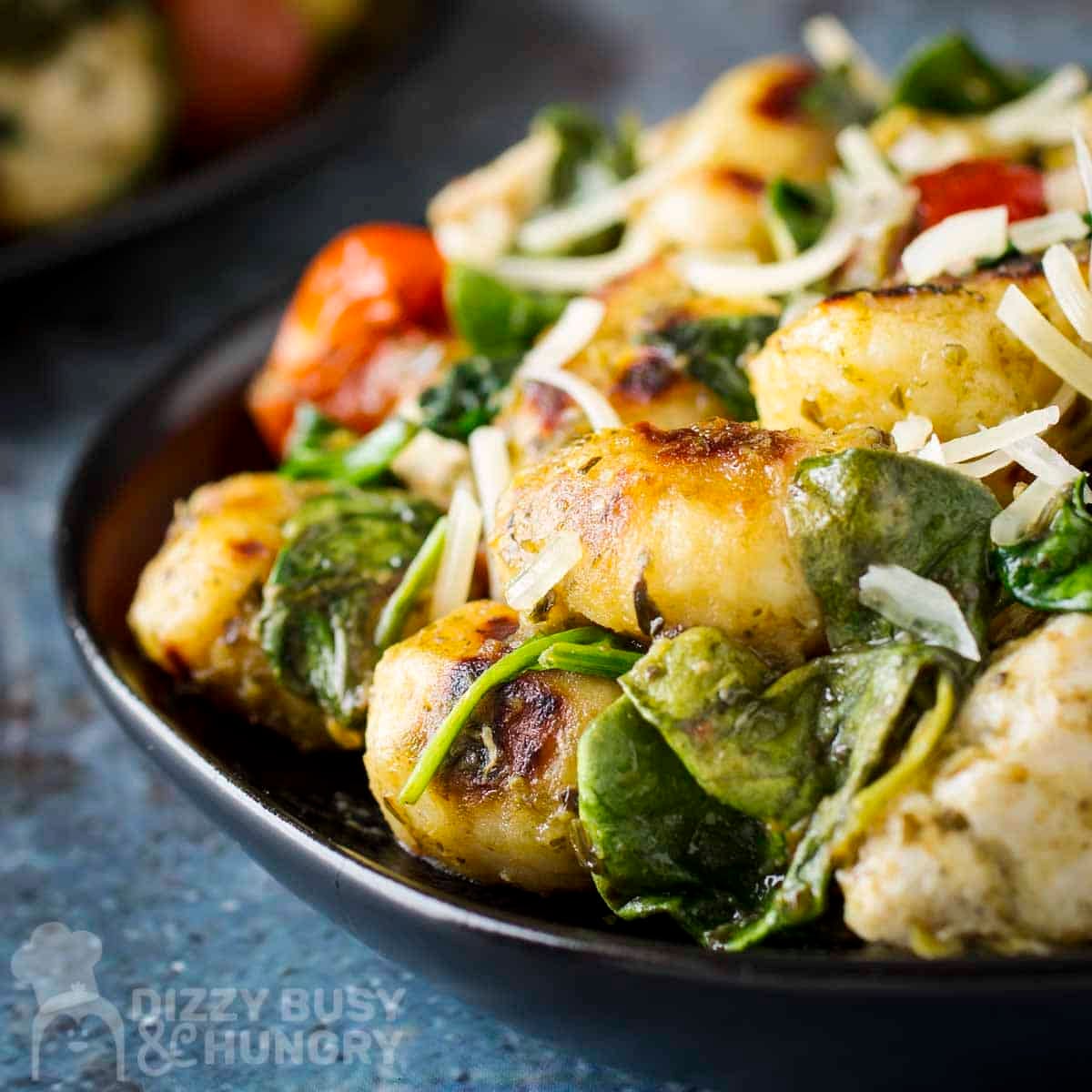 Side close up shot of pesto chicken and gnocchi sprinkled with shredded parmesan on a black plate.