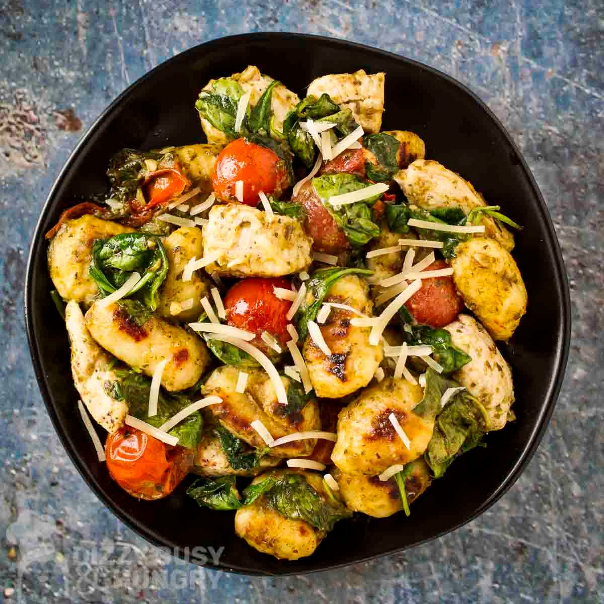 Overhead shot of pesto chicken and gnocchi sprinkled with shredded parmesan on a black plate.