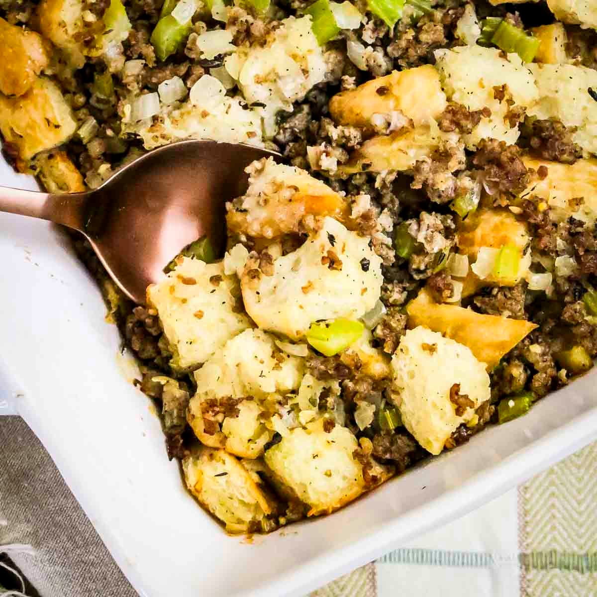 Close up shot of sage and sausage stuffing in a white baking dish with a spoon taking a scoop out.