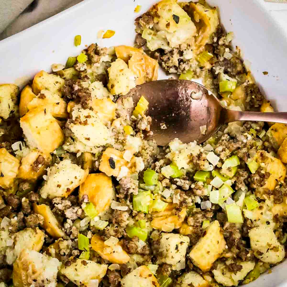 Close up shot of sage and sausage stuffing in a white baking dish with a spoon taking a scoop out.