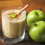Side shot of a clear glass with apple smoothie garnished with cinnamon, small apple chunks, and a cinnamon stick with two granny smith apples on the side.