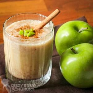 Side shot of a clear glass with apple smoothie garnished with cinnamon, small apple chunks, and a cinnamon stick with two granny smith apples on the side.