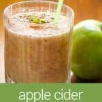Side shot of a clear glass with apple smoothie garnished with cinnamon, small apple chunks, and a straw with a granny smith apple on the side.