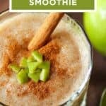 Close up shot of a clear glass with apple smoothie garnished with cinnamon, small apple chunks, and a cinnamon stick with a granny smith apple on the side.
