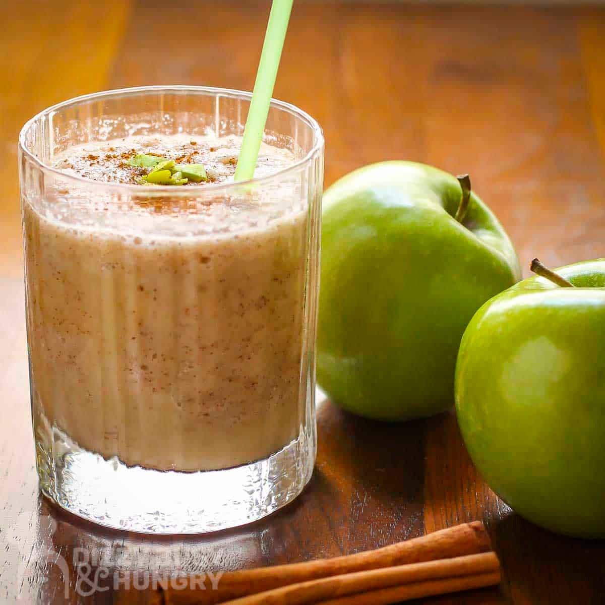 Side shot of a clear glass with apple smoothie garnished with cinnamon, small apple chunks, and a cinnamon stick with two granny smith apples on the side.