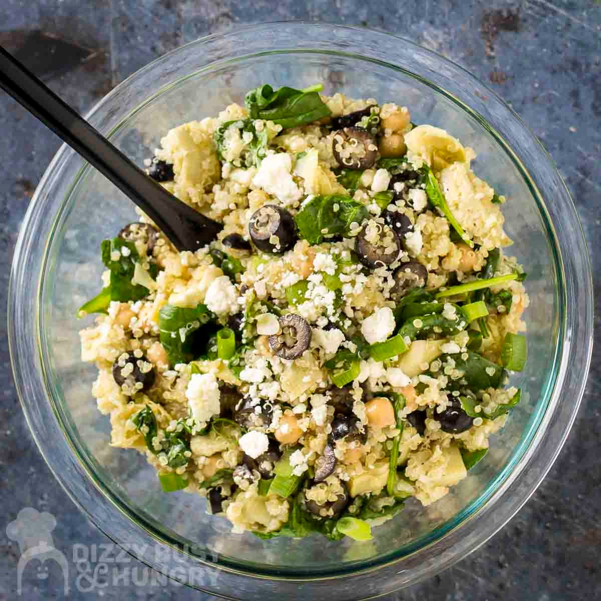 Overhead shot of quinoa salad in a glass bowl with a black spoon on a blue surface.