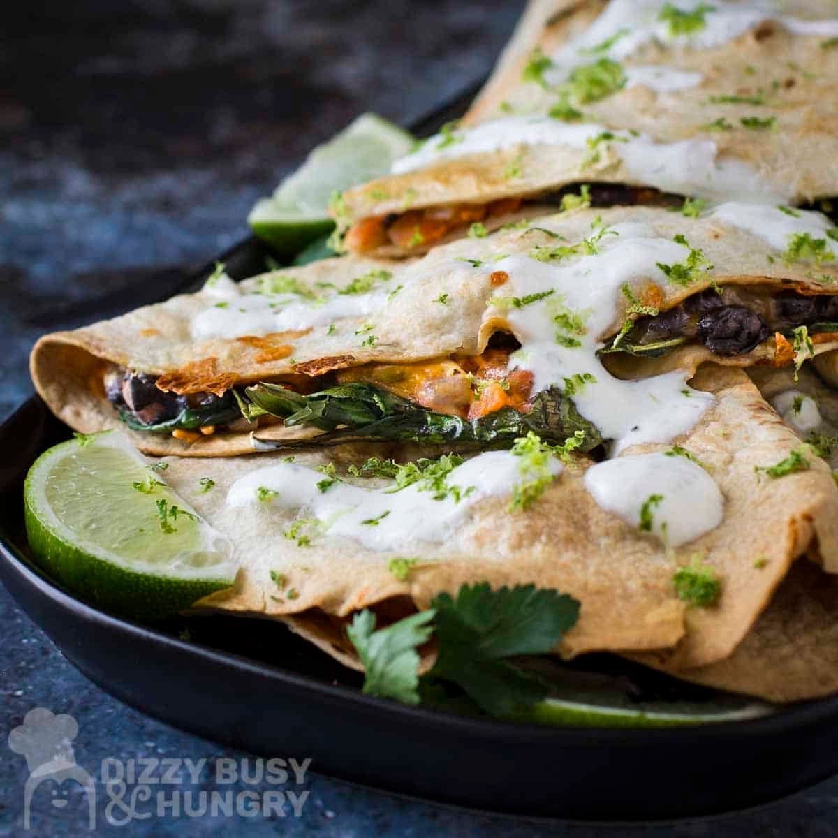 Side shot of multiple bean and cheese quesadillas garnished with fresh herbs and crema on a black plate with a lime wedge on the side.