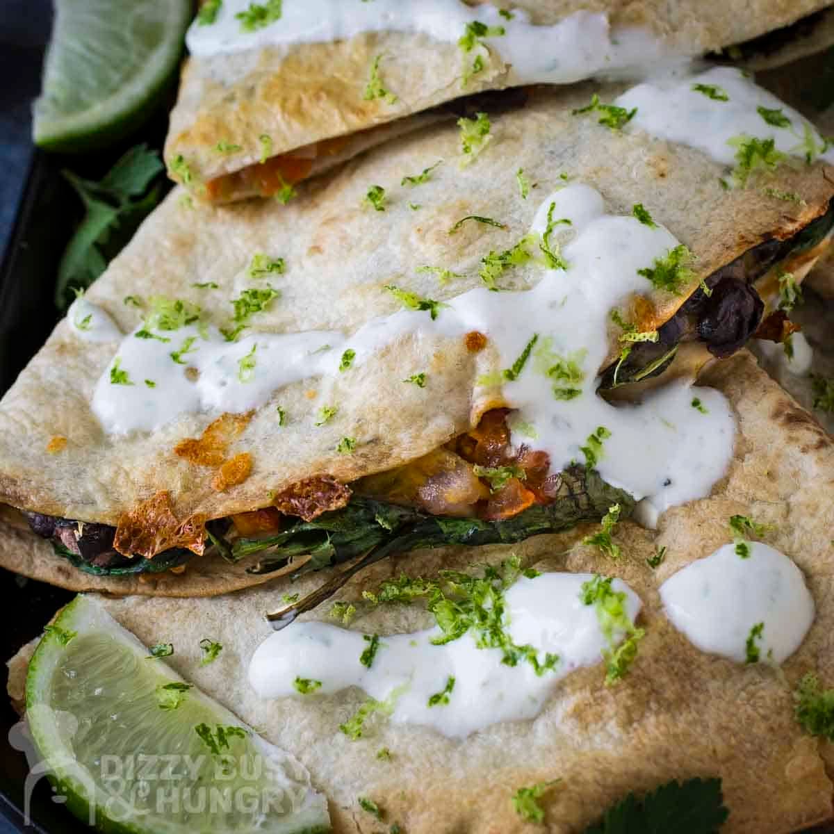 Overhead shot of multiple bean and cheese quesadillas garnished with fresh herbs and crema on a black plate with a lime wedge on the side.