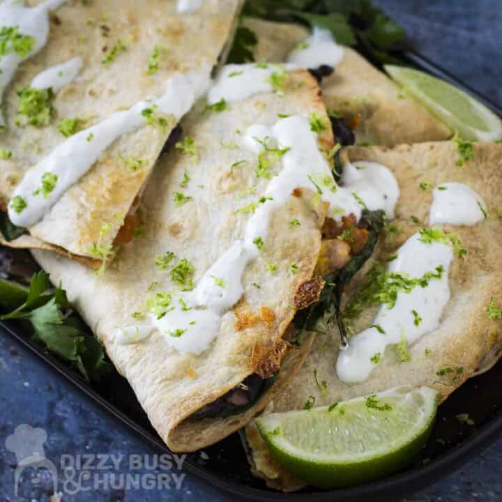 Side shot of multiple bean and cheese quesadillas garnished with fresh herbs and crema on a black plate with a lime wedge on the side.