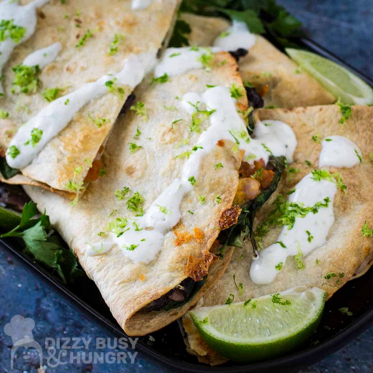 Side shot of multiple bean and cheese quesadillas garnished with fresh herbs and crema on a black plate with a lime wedge on the side.