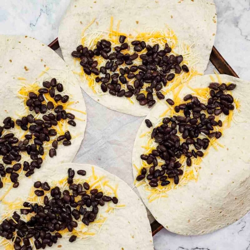 Overhead view of process shot 1 - place each tortilla on a baking sheet, draping half over the edge so that they all fit. Distribute about half of each of the cheese over the tortillas, then distribute all of the black beans.