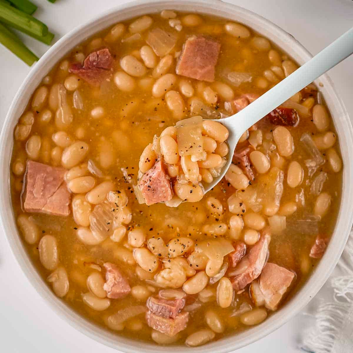 Overhead view of ham and bean soup in a white bowl with a spoon holding up a scoop on a white surface.
