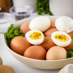 Side view of multiple boiled eggs with three eggs peeled and cut in half on top in a white bowl with fresh herbs on the sides.