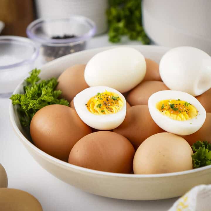 Side view of multiple boiled eggs with three eggs peeled and cut in half on top in a white bowl with fresh herbs on the sides.