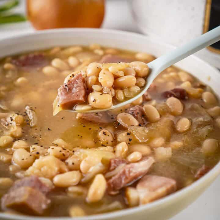 Side view of ham and bean soup in a white bowl with a spoon holding up a scoop on a white surface.
