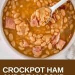 Overhead view of ham and bean soup in a white bowl with a spoon holding up a scoop on a white surface.
