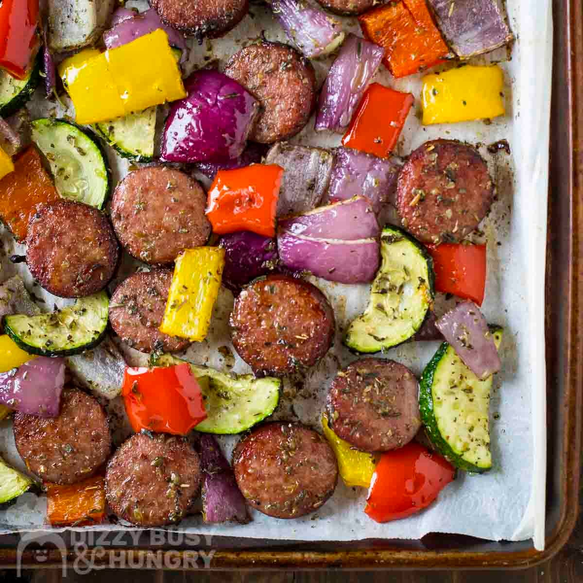 Close up overhead view of kielbasa and veggies on a parchment paper-lined baking sheet.
