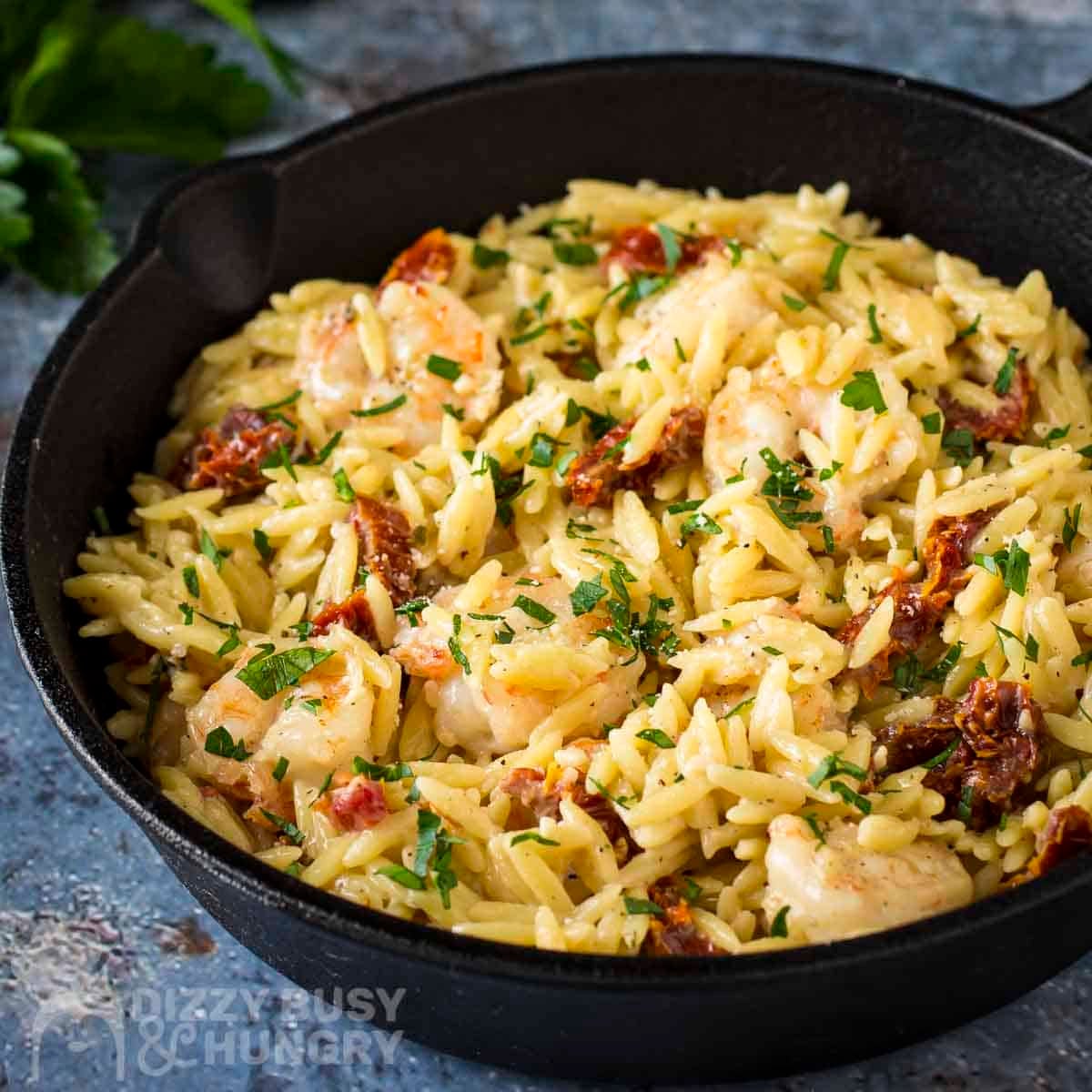 Side shot of shrimp and orzo garnished with fresh herbs in a cast iron skillet on a blue surface with fresh herbs in the background.