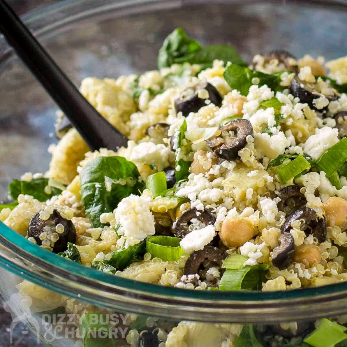 Side close up shot of quinoa salad in a glass bowl with a black spoon on a blue surface.
