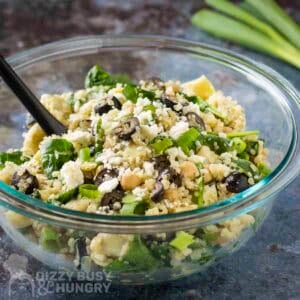 Side shot of quinoa salad in a glass bowl with a black spoon on a blue surface with scallions in the background.