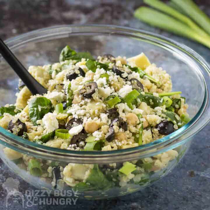 Side shot of quinoa salad in a glass bowl with a black spoon on a blue surface with scallions in the background.