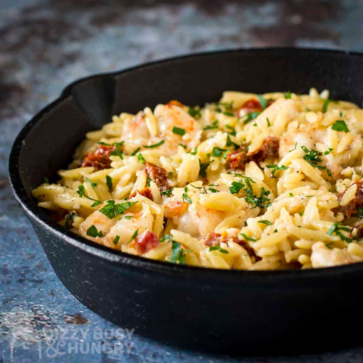 Side shot of shrimp and orzo garnished with fresh herbs in a cast iron skillet on a blue surface with fresh herbs in the background.