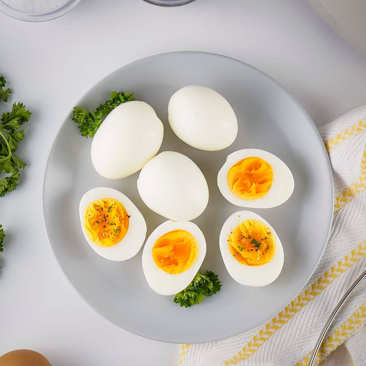 Overhead view of multiple peeled boiled eggs, some cut in half, on a white plate with fresh herbs on the side.