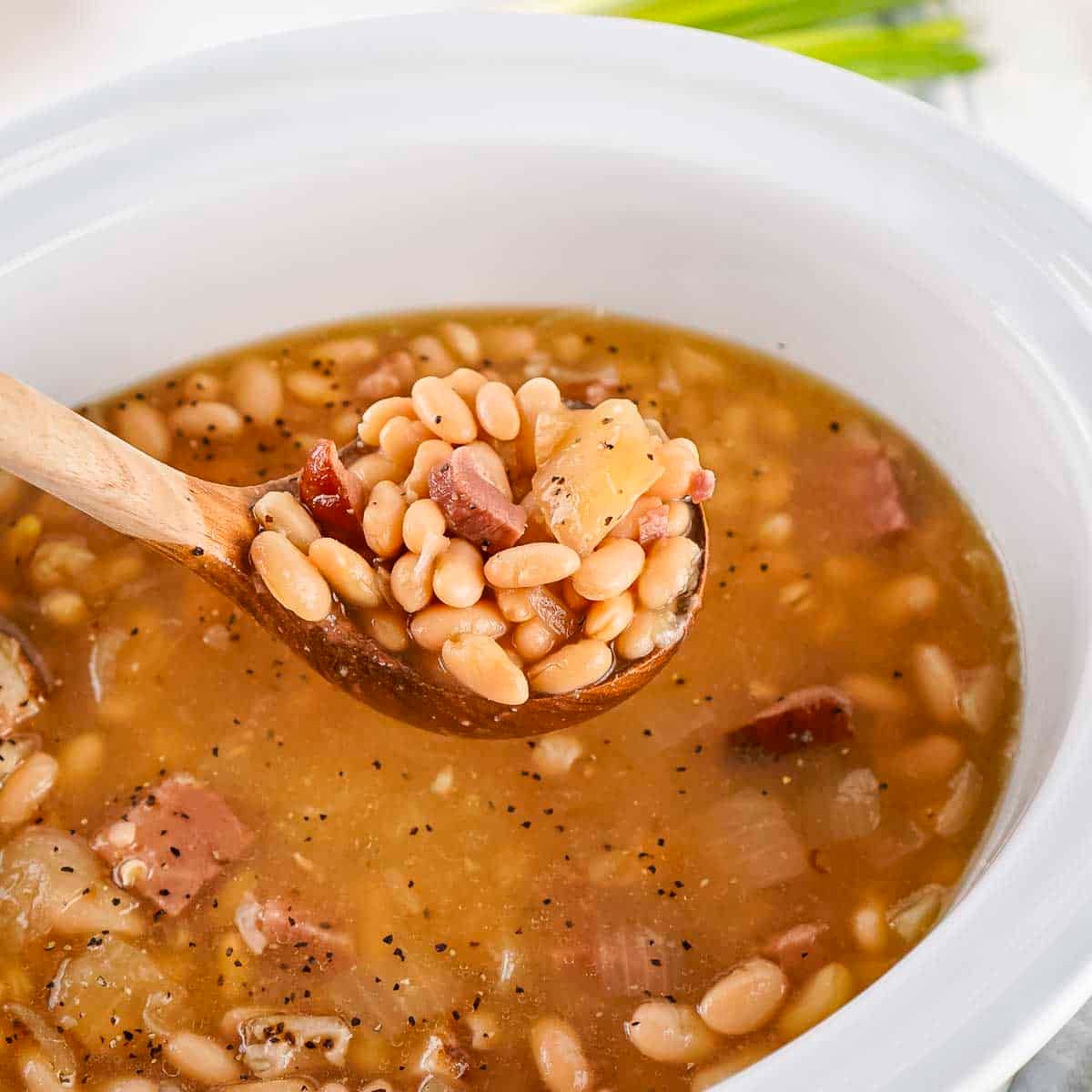Side view of ham and bean soup in a white crock pot with a spoon holding up a scoop on a white surface.