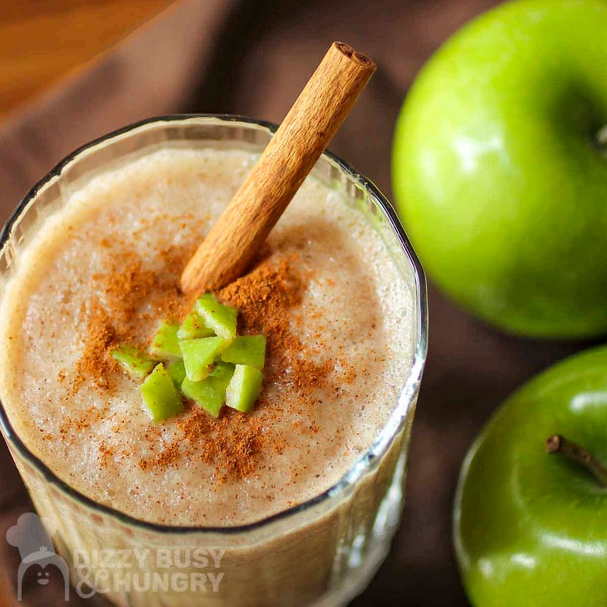 Overhead close up shot of a clear glass with apple smoothie garnished with cinnamon, small apple chunks, and a cinnamon stick with two granny smith apples on the side.