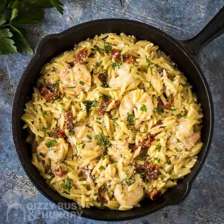 Overhead shot of shrimp and orzo garnished with fresh herbs in a cast iron skillet on a blue surface with fresh herbs in the background.