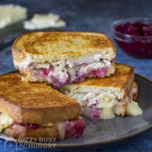 Side shot of three halves of turkey brie grilled cheese stacked on a black speckled plate with sliced brie and cranberry sauce in the background.