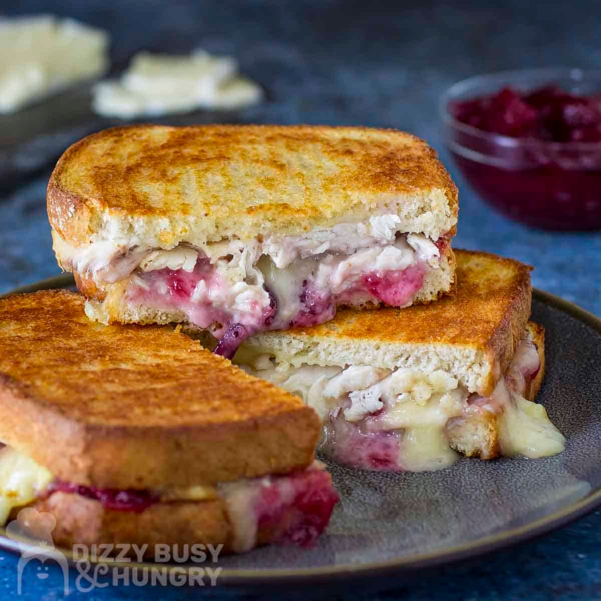Side shot of three halves of turkey brie grilled cheese stacked on a black speckled plate with sliced brie and cranberry sauce in the background.