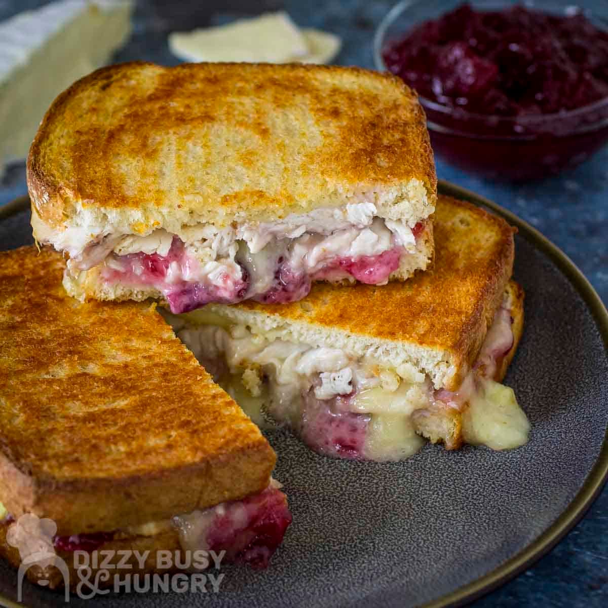 Side shot of three halves of turkey brie grilled cheese stacked on a black speckled plate with sliced brie and cranberry sauce in the background.