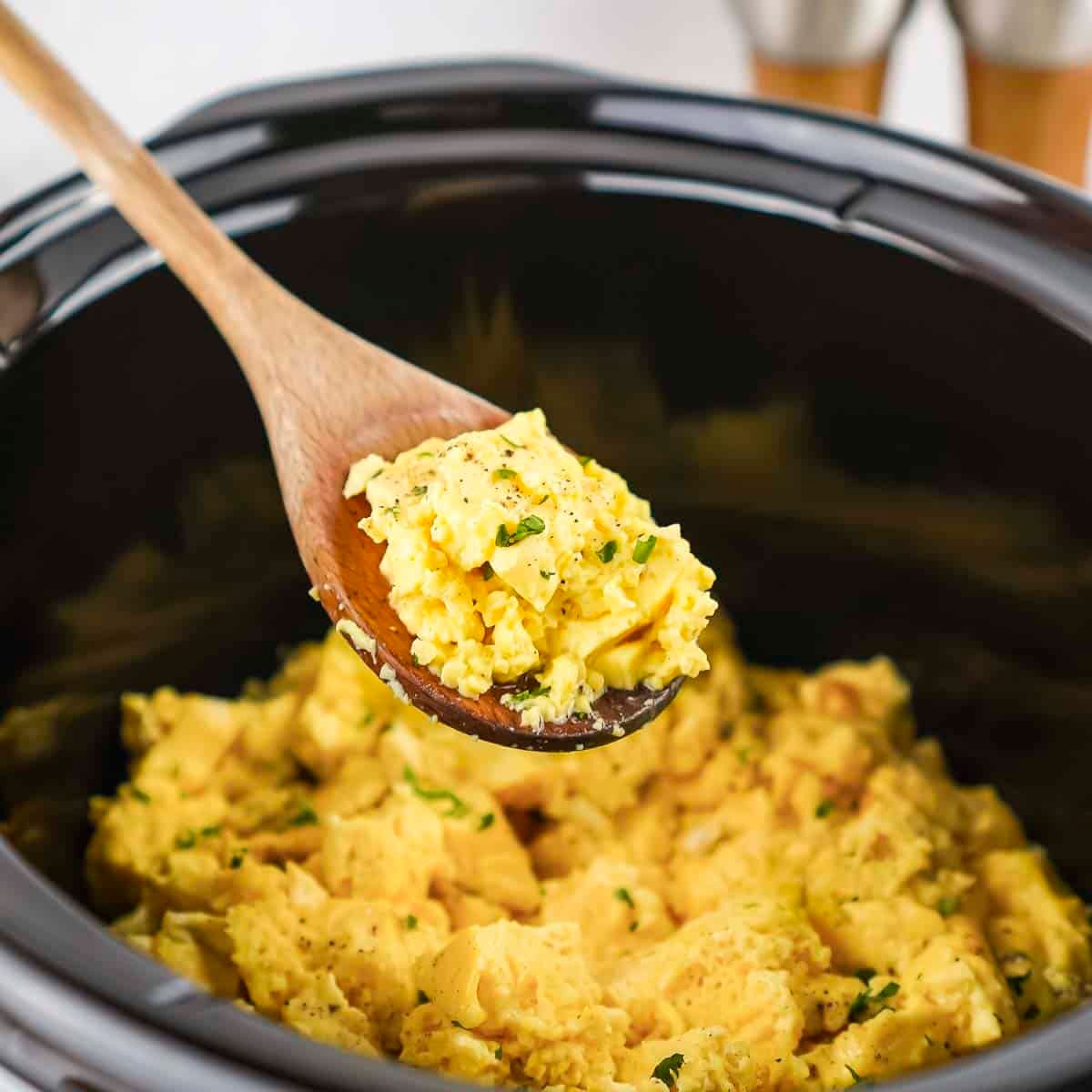 Side shot of a wooden spoon holding a scoop of eggs over a black crockpot bowl with scrambled eggs garnished with fresh herbs.