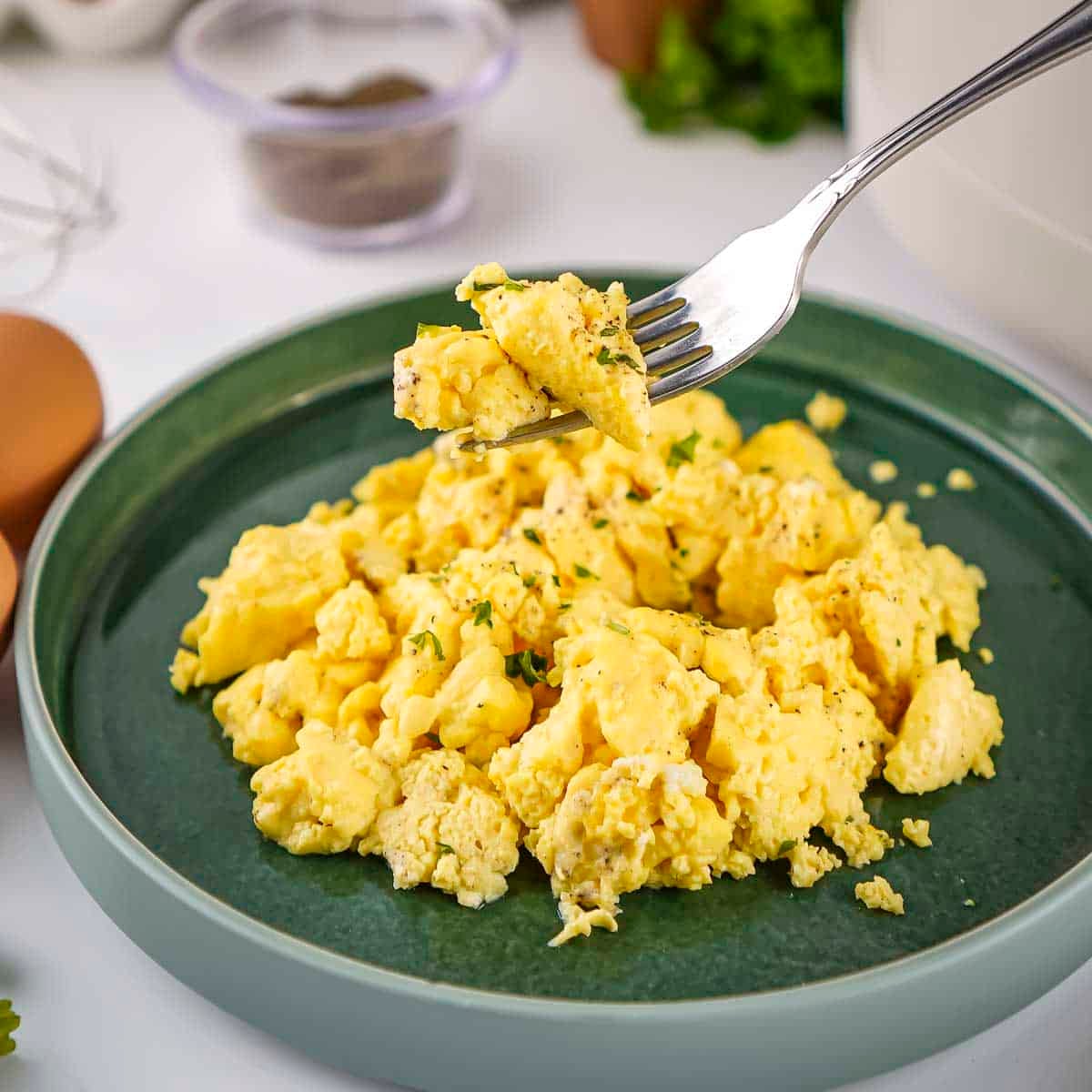 Side shot of a fork holding up a bite of eggs over a sage green plate of scrambled eggs garnished with fresh herbs.