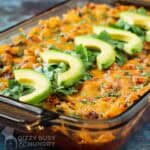 Side angled shot of dorito casserole in pan lined with chopped cilantro and slices of avocado.