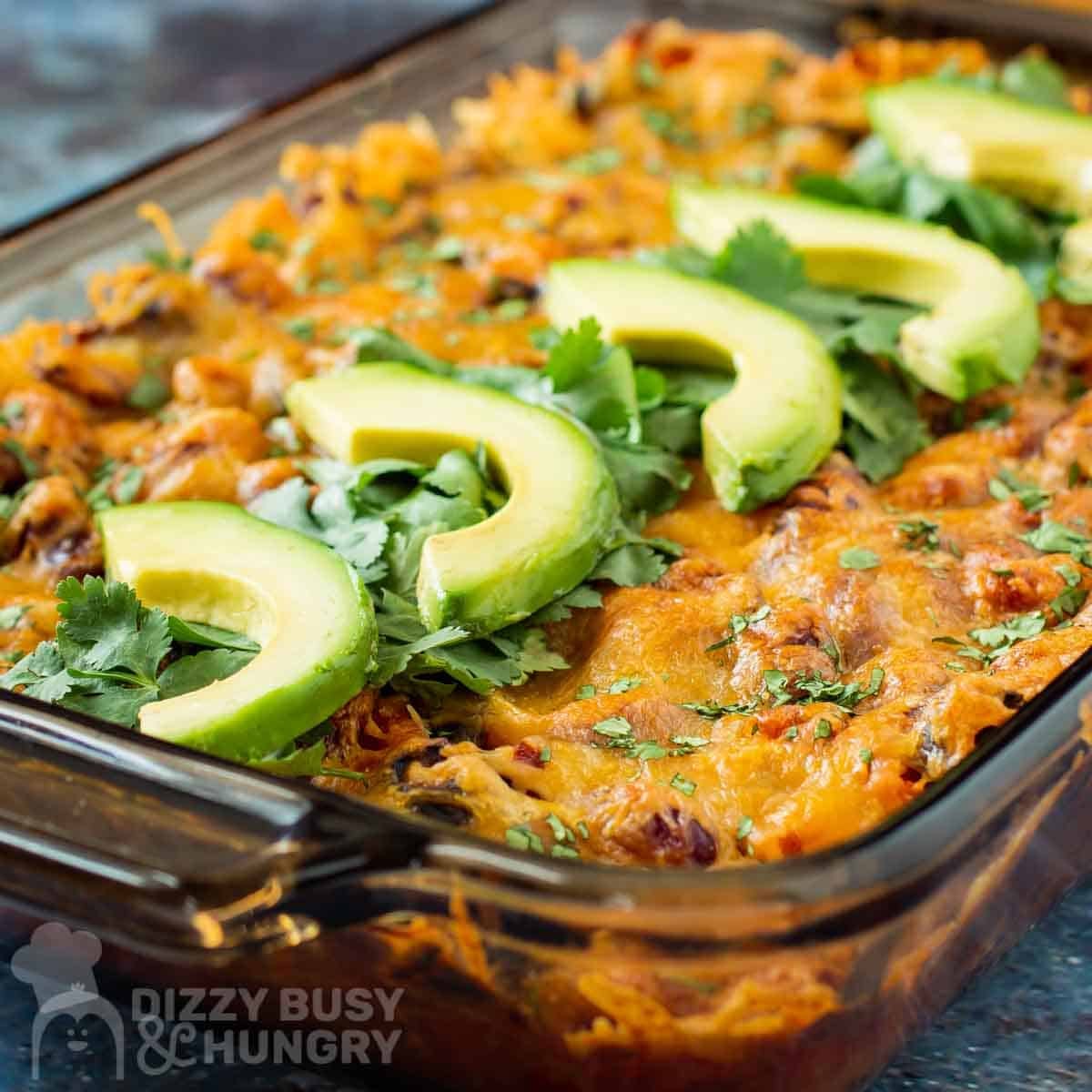 Side angled shot of dorito casserole in pan lined with chopped cilantro and slices of avocado.